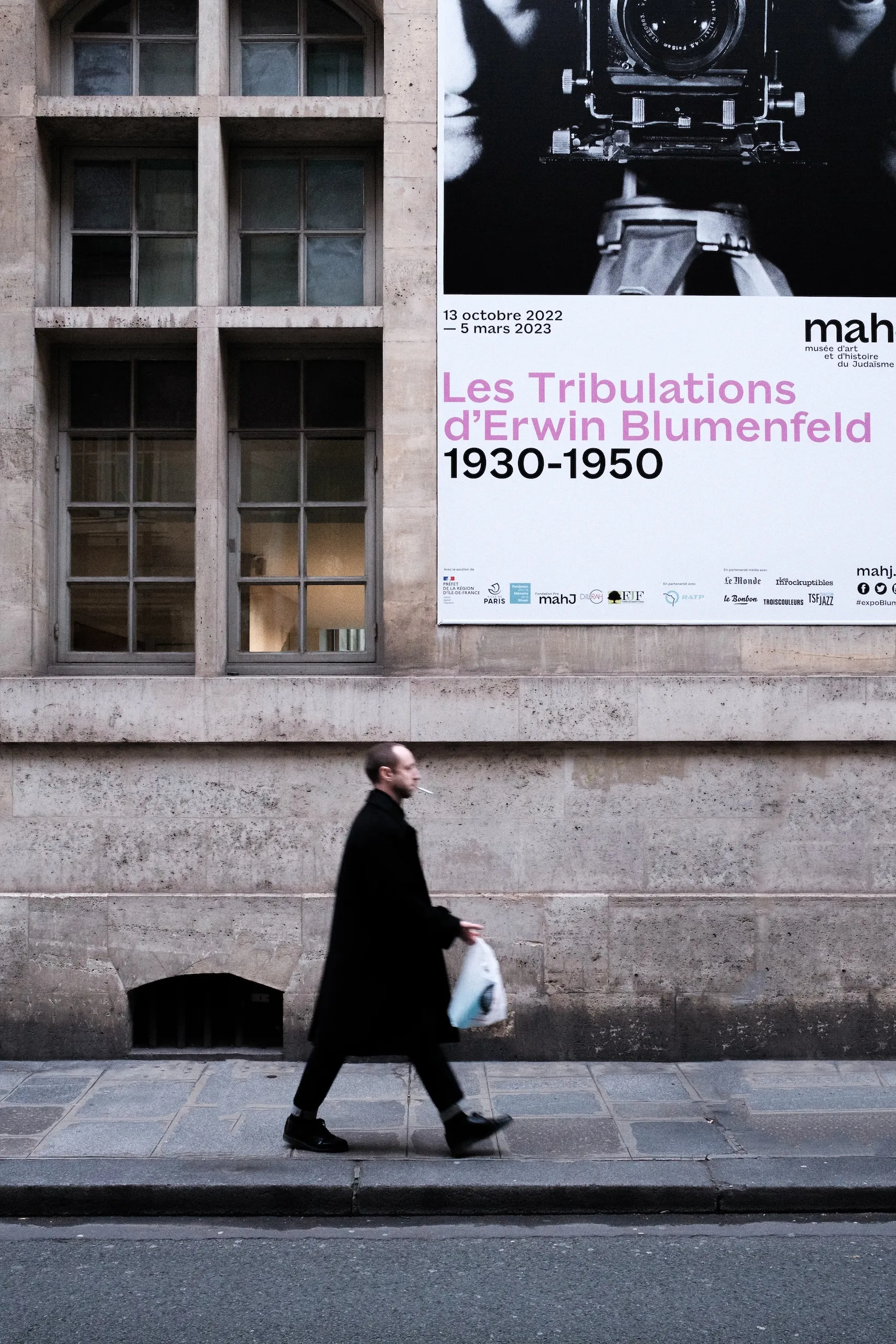 A man in a dark coat walking past an exhibition poster.